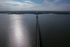 imagem 9, local: Aparecida do Tabuado-MS. Imagem aérea, a câmera aponta para frente. Na imagem vemos uma ponte sobre o rio Paraná, ela começa na parte inferior do quadro e sobe em direção a parte superior do quadro. Nesse ponto o rio tem 3 quilômetros de largura. No lado esquerdo do quadro o reflexo sol aparece na água, o lado direito está mais escuro. No horizonte vemos muitas nuvens espalhadas, o céu está azul claro. Nas margens a maior parte da área é desmatada, aparecem poucas árvores, há muitas áreas dedicadas a agricultura e pecuária.