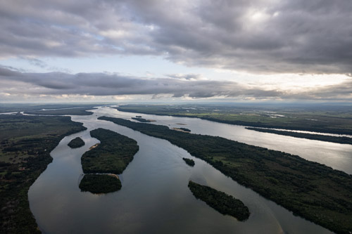 imagem 38, local: Rosana-SP.  Imagem aérea, a câmera aponta para o horizonte. Vemos o rio Paraná em sua extensão, o céu encoberto de nuvens, o sol se pondo do lado direito, a imagem é escura. O horizonte divide o quadro em duas partes iguais. Nesse ponto aparecem várias pequenas ilhas e extensões de terra na parte central do rio.  As ilhas são cobertas de mata e dividem o rio criando caminhos. O rio ocupa toda a parte inferior do quadro e segue em direção ao horizonte. Nesse ponto o rio tem 3 quilômetros e 700 metros de largura.