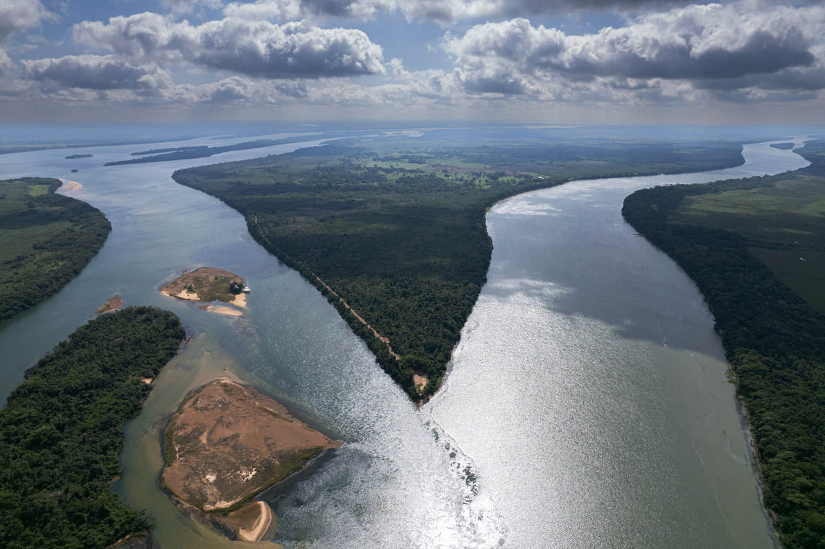 imagem 1, local: Marilena-PR. Encontro das águas dos rios Paraná à esquerda  e Paranapanema à direita. Imagem aérea, a câmera está a aproximadamente 500 metros de altura e aponta para frente. Esse é o ponto onde nasce o rio Paraná. Na imagem vemos o ponto de encontro dos rios Paraná à esquerda e Paranapanema à direita, as águas descem de cima para baixo no formato da letra ve, se encontram na parte inferior do quadro formando o rio Paraná. O sol reflete no ponto de encontro das águas dos rios, nuvens brancas projetam focos de sombras espalhados pela imagem. No centro da imagem, no ponto de encontro das águas temos o marco zero do Estado de São Paulo, é o local onde começa o estado, é a ponta esquerda do mapa do estado. As margens e o centro da imagem estão cobertas por matas e áreas verdes.