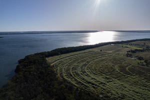 imagem 26, local: Castilho-SP. Imagem aérea, a câmera aponta para frente. Na imagem vemos uma fazenda as margens do rio Paraná, o rio reflete os raios solares. O céu está azul sem nuvens. No lado direito do quadro vemos uma fazenda com plantações não identificadas. A mata e o rio ocupam o canto inferior esquerdo, acompanham o rio fazendo uma curva em direção ao lado superior direito do quadro.