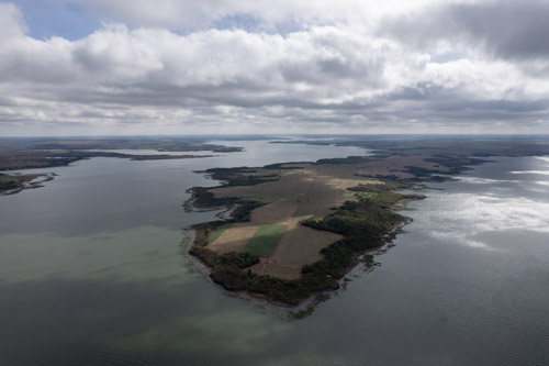 imagem 24: Local: Carneirinho-MG. Vista aérea do encontro dos Rios Parnaíba, à esquerda e Grande, à direita. Ponto onde nasce o rio Paraná. A câmera aponta para o horizonte, o drone está a 500 metros de altura. Ao fundo no centro da imagem temos uma faixa estreita de terra com florestas nas margens e terras preparadas para agricultura no centro. O céu está encoberto de nuvens brancas, que criam sombras sobre os rios. As nuvens estão bem próximas da câmera. No fundo à direita, o brilho do sol aparece refletido no rio Grande. A largura nesse ponto do rio é de quatro quilômetros, o drone estava a dois quilômetros da margem direita.