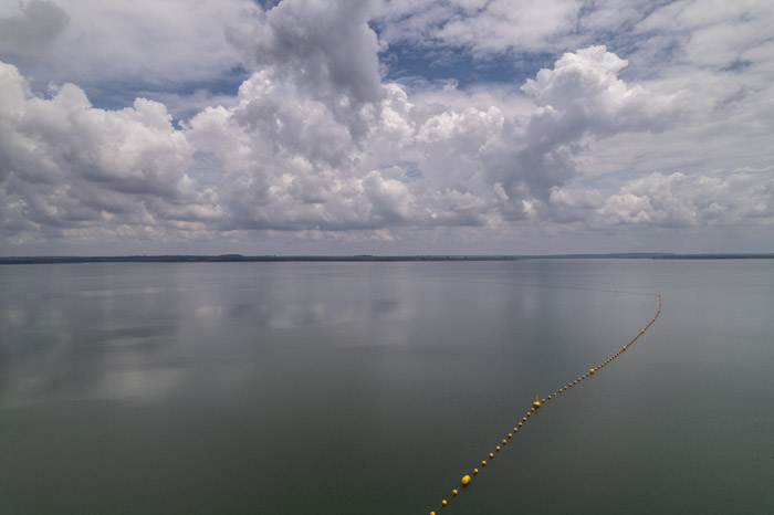 imagem 27, local: Ilha Solteira-SP. Imagem aérea, o drone está a 200 metros de altura, a câmera aponta para frente. Na imagem vemos o céu azul com grandes nuvens brancas, elas parecem estar baixas. O rio de águas verdes reflete as nuvens e o céu. No meio da parte inferior do quadro, sai uma corda com bóias amarelas, elas são redondas e estão conectadas a essa corda, elas sobem em direção ao horizonte no lado direito, depois continuam para o lado esquerdo. São bóias de segurança da hidroelétrica de Ilha Solteira.