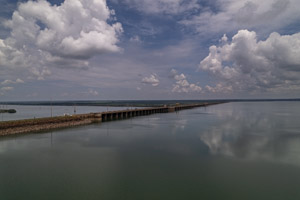 imagem 25, local: Ilha Solteira-SP. Imagem aérea, câmera aponta para frente. Na imagem vemos a Usina Hidroelétrica de Ilha Solteira, e o céu está azul, com grandes nuvens brancas refletidas nas águas do rio. A usina, uma estrutura em concreto, aparece numa diagonal que sai do lado esquerdo para o direito. No horizonte uma faixa escura divide o quadro em duas partes.