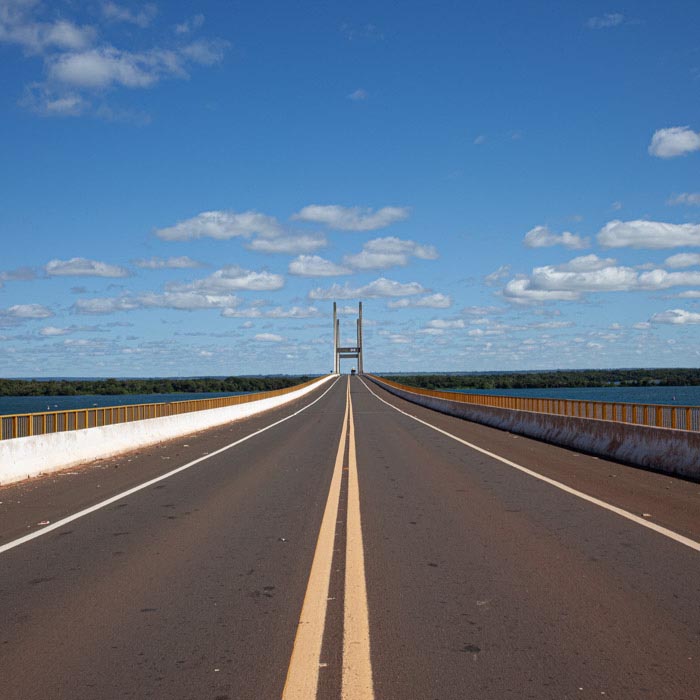 imagem 22, local: - Paulicéia-SP. Imagem feita de cima da ponte no meio da pista, a câmera aponta para frente. Na imagem vemos a estrada, um caminhão bem ao fundo, nas laterais o rio Paraná. O céu está azul com muitas nuvens brancas espalhadas. Do meio do quadro saem duas as faixas amarelas impressas na pista em direção ao lado superior do quadro, ao fundo uma estrutura de sustentação da ponte, vemos duas colunas grandes e uma terceira unindo as duas pelo meio. Na outra margem, a linha do horizonte é preenchida por uma mata.