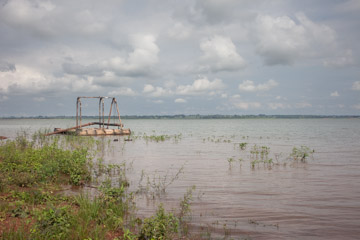 imagem 34, local: Rubinéia-SP. Imagem fotográfica, a câmera aponta para frente. Na imagem vemos o rio Paraná, e ao fundo, no lado esquerdo, uma estrutura flutuante utilizada para extração de areia. O céu está encoberto por diversas nuvens, e no primeiro plano do lado esquerdo, aparecem plantas rasteiras e aquáticas, na cor verde. O canto inferior direito é ocupado pela água. Na estrutura flutuante aparece um cano utilizado para sugar a areia do rio para fora. No horizonte distante, uma faixa escura formada por vegetação divide o quadro em duas partes iguais.