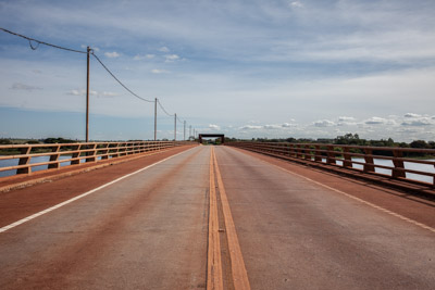 imagem 15, local: - Ilha Solteira-SP. Imagem feita de cima da ponte no meio da pista, a câmera aponta para frente. Na imagem vemos a estrada e nas laterais o rio Paraná. O céu está azul e parcialmente encoberto. Do meio do quadro saem duas faixas impressas na pista em direção ao lado superior do quadro, ao fundo uma estrutura de sustentação da ponte em formado de portal retangular. Há uma sequência de 7 postes no lado esquerdo da ponte, a pista está vazia.