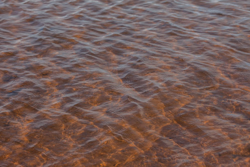 imagem 18, local: Rubinéia-SP. Imagem fotográfica, a câmera está a uma altura de 50 centímetros e aponta diagonalmente para baixo. Na imagem vemos a água do rio, na superfície reflexos do céu azul, no fundo a terra marrom alaranjada. Nessa imagem os raios solares iluminam o fundo do rio que está a uma profundidade de aproximadamente 20 centímetros. Na superfície, o sol de final de tarde ilumina de cima para baixo, deixando a parte de cima da foto com mais reflexos e a parte de baixo mais transparente e alaranjada. O movimento da água causado pela correnteza e pelo vento criam distorções na luz do sol que atinge o fundo do rio.