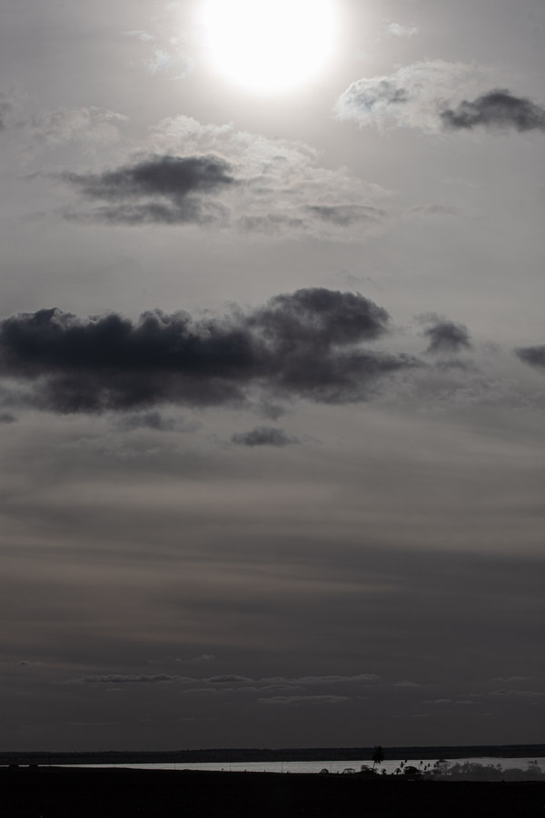 imagem 37, local: Rubinéia-SP. Imagem fotográfica na vertical, a câmera aponta para o horizonte. No topo da imagem vemos o brilho do sol, o céu está parcialmente encoberto por nuvens escuras. No rodapé do quadro, vemos o rio que está muito distante, aparece apenas uma pequena faixa prateada na horizontal. A câmera aponta diretamente para o sol que aparece quase inteiro na imagem. Para fotografar o sol, a câmera precisa cortar a entrada de luz, por isso, a imagem é escura.