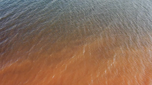 imagem 36, local: Santa Clara d’Oeste-SP. Imagem aérea em câmera lenta, a câmera aponta diagonalmente para baixo. A duração do video é de 35 segundos. Na imagem vemos as águas do rio Paraná, na parte de cima ela é verde, na parte de baixo do quadro a água é marrom. A correnteza, lentamente, movimenta a água da esquerda para a direita, com esse movimento, o brilho do sol refletido na água cria pequenos pontos prateados na superfície, que aparecem e desaparecem com o movimento das águas. A água do rio é verde; o marrom que aparece na imagem vem do barro que se solta da margem do rio.