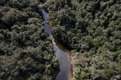 Imagem 9, local: Parque Estadual Nascentes do Paranapanema, Capão Bonito-SP. Imagem aérea de drone, a câmera aponta para baixo. Na imagem vemos o rio Paranapanema cercado pela mata. Nesse ponto o rio é estreito e raso, a água é cristalina de tom alaranjado, reflete nuvens e um pouco do azul do céu.