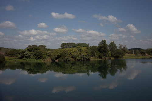 Imagem 34, local: Jacarezinho-PR. Imagem fotográfica, a câmera aponta para frente. Imagem feita de cima de uma ponte. Na margem do rio é possível ver algumas árvores de diferentes espécies e tons variados de verde. No canto esquerdo tem um guarda-sol e alguém parece estar pescando. O dia está ensolarado, o céu azul e as nuvens brancas aparecem refletidas no rio.  As nuvens refletidas na água formam um círculo oval com a imagem espelhada no rio.