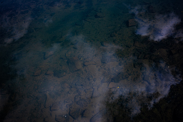 Imagem 38, local: Jacarezinho-PR. Imagem fotográfica, a foto foi tirada de cima de uma ponte, a câmera aponta para baixo. Na imagem vemos as pedras no fundo do rio e reflexos das nuvens. A imagem é escura, porém, a água cristalina permite ver as pedras no fundo do rio, algumas nuvens brancas, e na parte superior do quadro, o tom esverdeado da água que se mistura com a cor marrom das pedras. A imagem recebeu tratamento para realçar as cores e contraste, deixando os pretos mais escuros e realçando os brancos das nuvens.