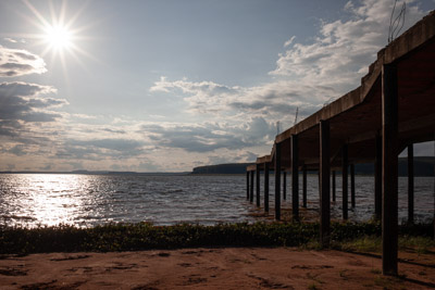 Imagem 40, local: Paranapanema-SP. Imagem fotográfica, a câmera aponta para frente. Do lado esquerdo da imagem vemos uma estrutura de concreto que se projeta para dentro do rio. Do lado direito, o sol aparece no céu e também refletido na água. O céu está parcialmente encoberto. Essa estrutura está abandonada, e é formada por uma laje de concreto sustentada por diversas colunas que se projeta para dentro do rio, tem aproximadamente três metros de altura, não é apropriada para atracar barcos, porque é muito alta. Curiosidade: A estrutura é mais baixa na parte oposta ao rio. É possível acessar a parte superior e observar a paisagem de cima, nesse ponto o rio tem uma largura aproximada de um quilometro e cem metros.