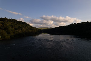 Imagem 42, local: Parque Natural do Dourado - Piraju-SP. Imagem aérea, a câmera aponta para o horizonte. Na imagem vemos o rio Paranapanema que preenche metade do quadro. Ao fundo, céu azul com algumas nuvens brancas. As margens são cobertas por uma mata fechada. É fim de tarde, a luz do sol ilumina a margem esquerda do rio, a mata da margem direita projeta sombras deixando as águas do rio escuras. Curiosidade: nesse ponto o rio tem uma largura aproximada de 150 metros.