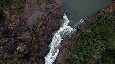 Imagem 29, local: Piraju-SP. Imagem aérea de vídeo, a câmera aponta para baixo. Na imagem vemos o rio estreito correndo sobre solo rochoso, vegetação verde no lado direto do quadro e rochas no lado esquerdo. Nesse ponto, o rio se afunila e forma um desnível, fazendo com que as águas, que correm de cima para baixo, ganhem velocidade, o atrito com as pedras produz uma espuma branca que se intensifica à medida que o rio passa nesse ponto. Num segundo momento o vídeo mostra uma imagem mais fechada desse mesmo trecho do rio, só que agora mais focado no movimento das águas e nas espumas que se formam.