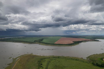 Imagem 46, local: Guaraci-SP. Imagem aérea de vídeo. A câmera aponta para o horizonte. A imagem mostra o rio Grande e suas margens, o céu com nuvens carregadas, e mais adiante, a chuva. Na parte inferior do quadro vemos a margem com vegetação verde e rasteira. Acima, na outra margem, ao redor do rio,  uma faixa de mata verde escura e lavouras de plantações espalhas pelo quadro. No horizonte, bem distante, vemos a massa cinzenta de chuva encobrindo o céu.