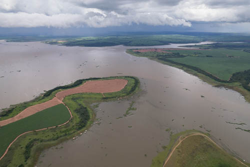 Imagem 39, local: Guaraci-SP. Imagem aérea, a câmera aponta para o horizonte. A fotografia mostra o rio Grande em sua extensão, grandes lavouras de plantações, casas flutuantes na margem esquerda do rio. O céu está encoberto por nuvens densas e escuras. Mais adiante, no horizonte, chove.