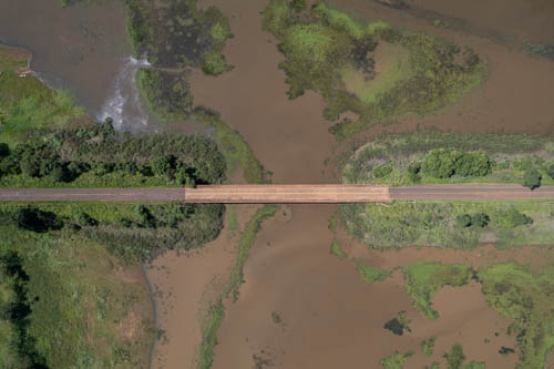 Imagem 27, local: Indiaporã-SP.  Imagem aérea, a câmera aponta para baixo. A fotografia mostra uma ponte no centro da imagem que liga duas estradas. O quadro é quase todo preenchido por áreas alagadas. Embaixo da ponte vemos a água do rio na cor marrom, ela se espalha nas áreas ao redor das estradas. No lado esquerdo aparecem algumas árvores e arbustos ao redor da estrada. Na parte superior esquerda aparecem dois barcos de pesca, na imagem parecem ser menor do que um grão de arroz.