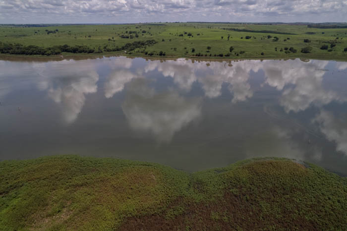 imagem 40, local: Iturama-MG. Imagem aérea, a câmera aponta para o horizonte. A imagem mostra o Rio Grande, suas margens e o céu azul de nuvens brancas refletido nas águas. No primeiro plano vemos uma das margens do rio coberta por vegetação. Acima, vemos o rio, e ao fundo, na outra margem, aparecem árvores distribuídas numa grande área verde de pastagem. As árvores são verdes e mais escuras que os pastos. O céu é azul, com nuvens de diferentes tamanhos que se espalham até o horizonte. As águas escuras do rio refletem o azul do céu e as grandes nuvens brancas.