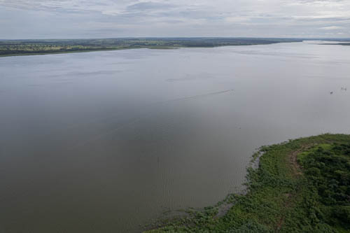 imagem 41, local: Mesópolis-SP. Imagem aérea, a câmera aponta para o horizonte. A imagem mostra o Rio Grande e suas margens. Nesse ponto o rio tem 2.500 metros de largura. No canto inferior direito, vemos parte da margem do rio, no horizonte vemos a outra margem. Na imagem, vemos a parte estreita do rio no lado direto, e a parte mais larga no lado esquerdo. A água é escura, é possível ver um barco de pesca subindo o rio na altura do meio do quadro, ele aparece na imagem menor que um grão de arroz. O céu está encoberto por uma massa de nuvens grandes.