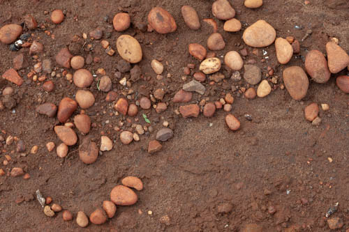 Imagem 14, local: Mesópolis-SP. Imagem fotográfica, a câmera está a 60 centímetros de altura do chão e aponta para baixo. Na imagem vemos várias pedras de rio, de diferentes tamanhos e cores, a maioria tem formato arredondado e a cor varia entre tons de marrom claro ao marrom avermelhado.