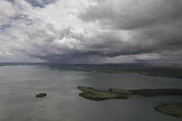 imagem 45, local:  Miguelópolis-SP. Imagem aérea de vídeo. A câmera aponta para o horizonte. O drone está à 335 metros de altura. Na imagem vemos o rio em sua extensão, ao fundo nuvens carregadas e uma massa cinza no ar, é a chuva que cai. No céu, as nuvens estão carregadas e escuras na maior parte. O rio de águas verdes apresenta grandes sombras projetadas pelas nuvens. A imagem é escura, a chuva se aproxima da direita para esquerda.