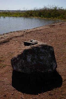 Imagem 15, local: Mesópolis-SP. Imagem fotográfica, a câmera aponta para frente. Na imagem vemos um par de sapatos em cima de uma pedra. À frente, a margem do rio e ao fundo uma vegetação baixa na linha do horizonte. A pedra tem aproximadamente 50 centímetros de altura, os sapatos estão com as solas viradas para cima. O chão é de cor marrom avermelhada, o céu está azul e sem nuvens. O sol ilumina o lado oposto da pedra e cria uma sombra escura que se projeta no chão se integrando ao formato da pedra.