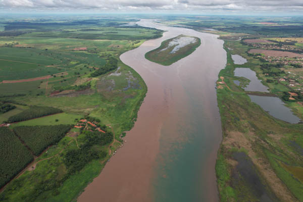 imagem 2, local: Planura-MG. Imagem aérea, a câmera aponta diagonalmente para baixo. Vemos o rio na sua extensão, mais a frente uma ilha. Uma faixa estreita de céu encoberto por nuvens. O horizonte, distante, se funde com o céu. Na margem direita, aparecem 3 lagos paralelos ao rio e a cidade de Planura-MG. Na margem esquerda, muitas áreas verdes, fazendas, plantações e algumas áreas com matas. O rio tem uma coloração marrom com uma mancha verde no lado inferior direito.