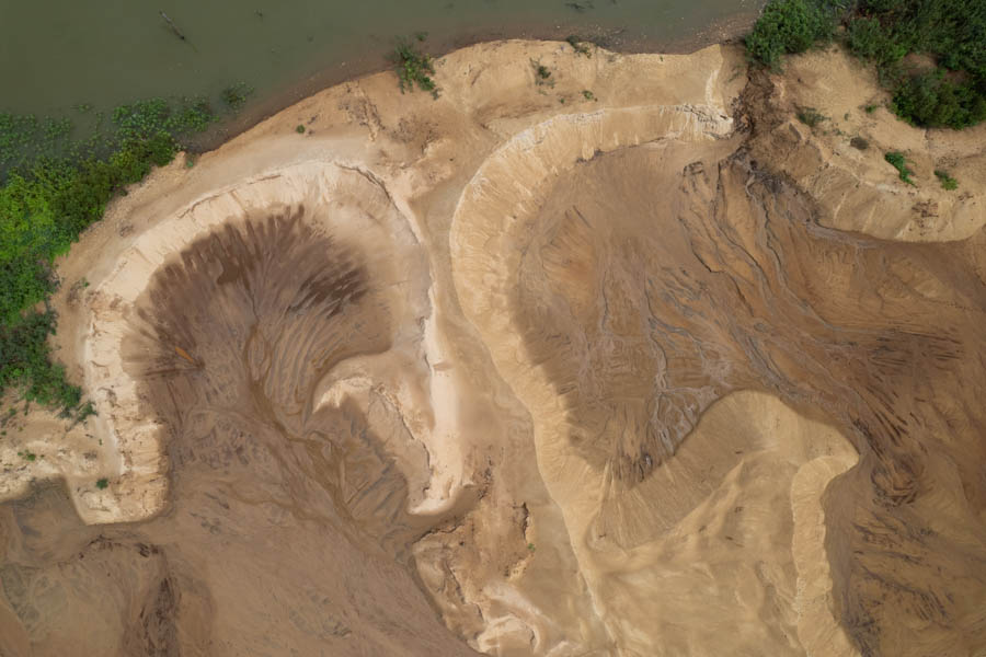 imagem 6, local: Populina-SP. Imagem aérea, a câmera aponta para baixo. A foto valoriza as várias texturas e formatos criados pela extração de areia do local. Na imagem vemos um pequeno pedaço do rio na parte superior do lado esquerdo, algumas plantas aquáticas e arbustos. O restante da imagem é preenchido por terra e areia. A coloração varia entre o marrom mais claro ao mais escuro. A movimentação da água na superfície do solo cria fissuras na terra, são os percursos por onde a água tenta encontrar caminhos.