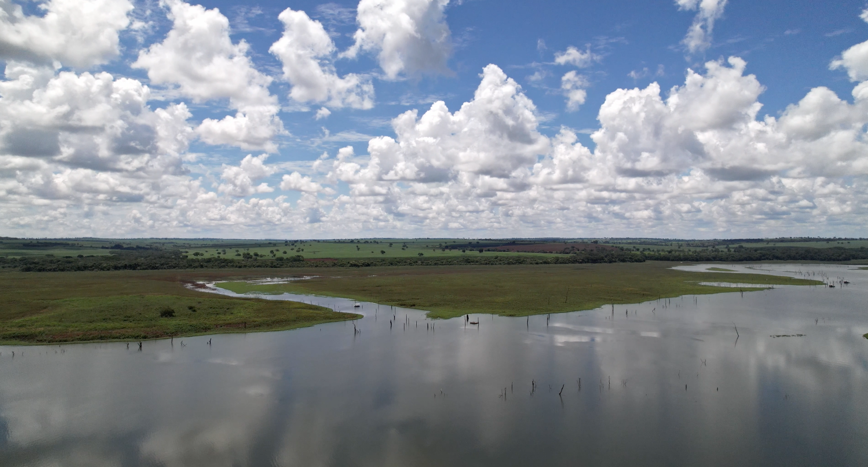 imagem 22, local: Iturama-MG. Imagem aérea de vídeo, a câmera aponta para o horizonte, o drone faz um movimento giratório de 360 graus em seu eixo, o tempo de duração é de 50 segundos. O drone foi posicionado no meio do rio a uma altura de aproximadamente 200 metros. A imagem giratória mostra o céu azul com nuvens brancas e o rio com os seus leitos e extensões. O horizonte divide o quadro ao meio. A água do rio reflete o azul do céu e as nuvens brancas. O que se vê ao redor são pastos, fazendas e poucas árvores.