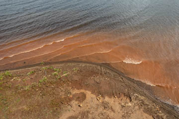 Imagem 18, local: Santa Clara D'Oeste-SP. Imagem aérea, a câmera aponta diagonalmente para baixo. O drone está baixo. Na parte superior aparece o rio Grande, na parte inferior a margem do rio. A terra sai do canto inferior direito e faz um arco em direção à lateral esquerda do quadro. A terra está parcialmente molhada e aparecem algumas plantas rasteiras no lado esquerdo da imagem. Da esquerda para a direita, o vento agita a superfície da água e cria pequenas ondas, as ondas se desmancham ao tocar a terra.